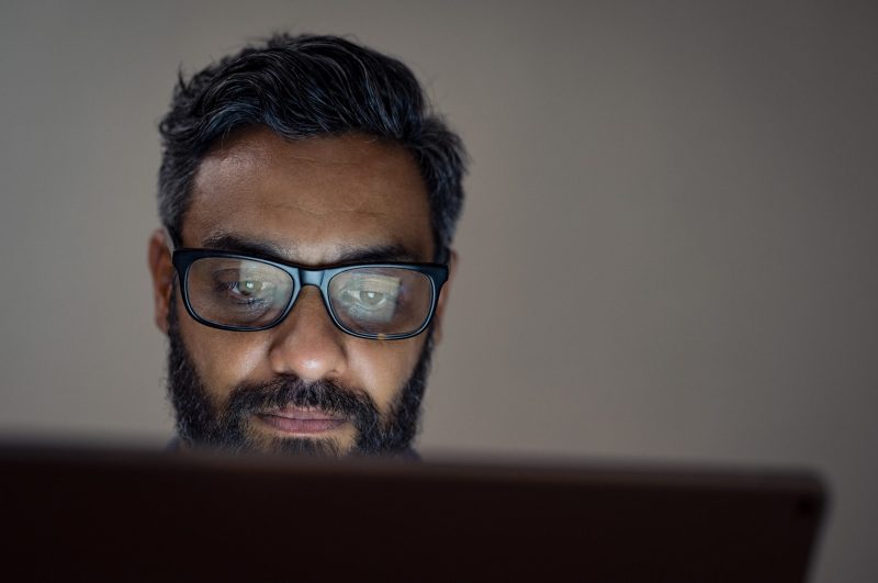 Mature man wearing eyeglasses sitting and using laptop in a dark office.